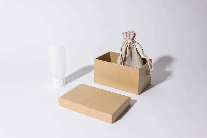 Empaque de cartón kraft, bolsa de lino y tubo de producto sobre fondo claro.
